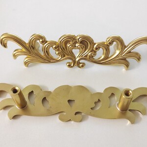 May include: A pair of gold-toned metal drawer pulls with an ornate, floral design. The pulls have a decorative, scroll-like shape with intricate details.