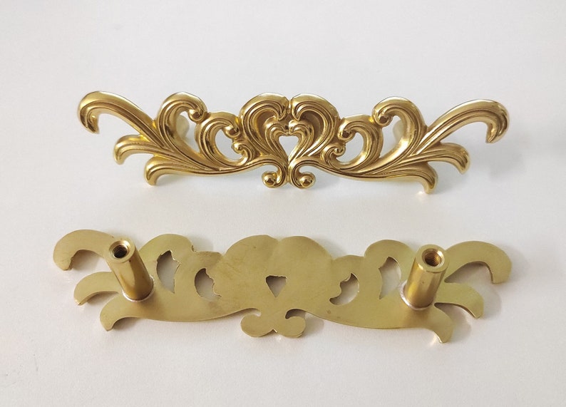 May include: A pair of gold-toned metal drawer pulls with an ornate, floral design. The pulls have a decorative, scroll-like shape with intricate details.