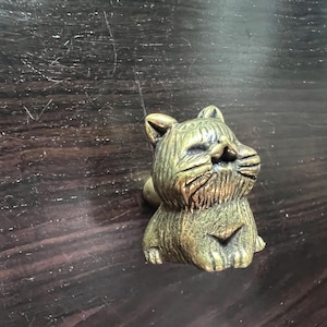 May include: A small, detailed brass cat figurine with a textured surface. The cat is in a seated position with its eyes closed, and has a small tail. The figurine is on a dark brown wooden surface.