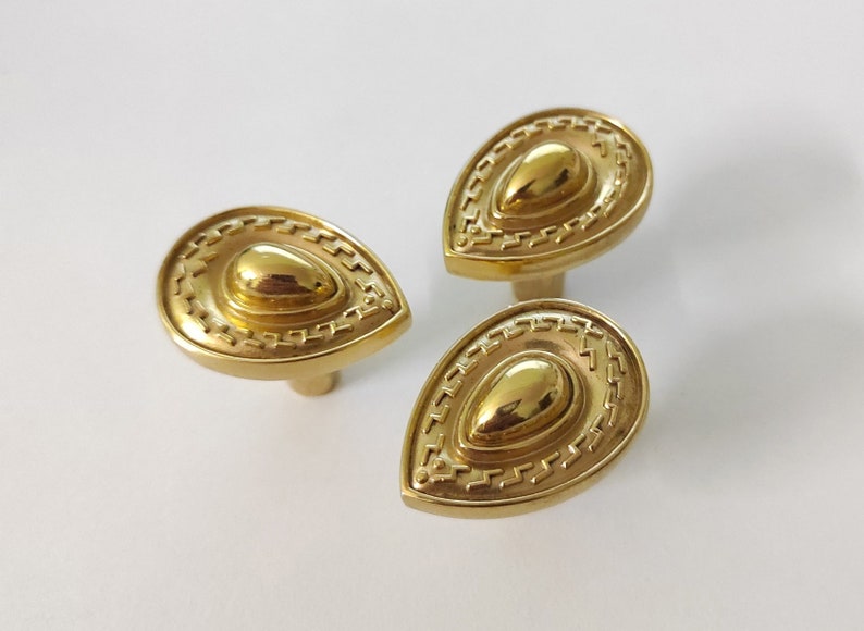 May include: Three gold-toned teardrop-shaped cabinet knobs with a decorative, raised, concentric circle design.
