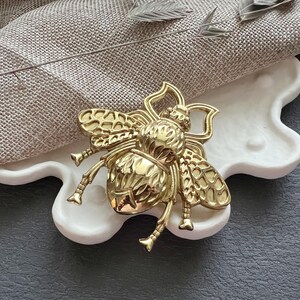 Puede incluir: Un objeto decorativo en forma de abeja de color dorado con detalles intrincados. La abeja está sentada en una bandeja de cerámica blanca con una forma irregular.