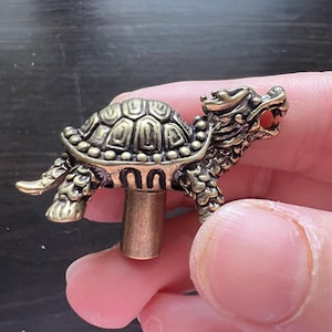 Könnte beinhalten: Detaillierte Schildkrötenfigur aus Messing mit einem drachenartigen Kopf. Die Schildkröte hat einen strukturierten Panzer und ist auf einer Messingstange montiert. Die Figur ist etwa 5 cm lang.