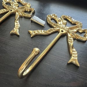 May include: Two gold-coloured metal wall hooks with a bow design. The hooks are made of a shiny, reflective metal and include mounting hardware. The hooks are designed to be mounted on a wall for hanging items.