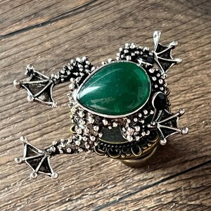 以下が含まれることがあります： カエル型の装飾的なボトルストッパー。ストッパーは、銀色の金属フレームにセットされた、大きな涙型の緑色の宝石が特徴です。カエルは、詳細な足と質感のある体、黒い宝石の目を持っています。