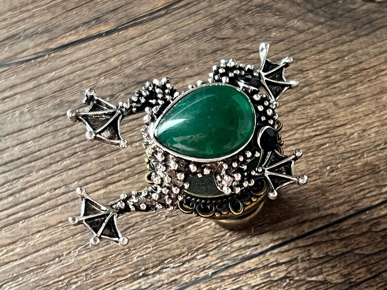 以下が含まれることがあります： カエル型の装飾的なボトルストッパー。ストッパーは、銀色の金属フレームにセットされた、大きな涙型の緑色の宝石が特徴です。カエルは、詳細な足と質感のある体、黒い宝石の目を持っています。