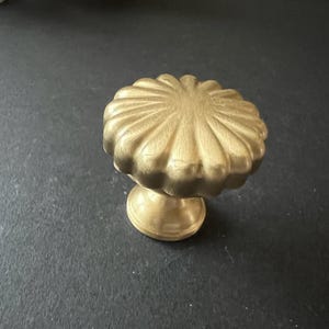 Puede incluir: Un pomo de armario dorado con un dise&ntilde;o acanalado y festoneado. El pomo tiene una base redonda y una parte superior circular elevada.