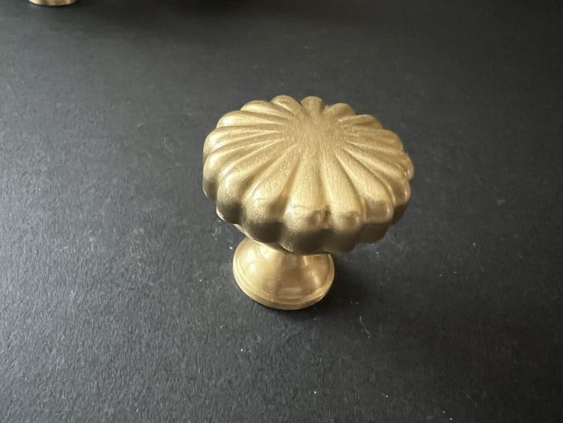 Puede incluir: Un pomo de armario dorado con un dise&ntilde;o acanalado y festoneado. El pomo tiene una base redonda y una parte superior circular elevada.