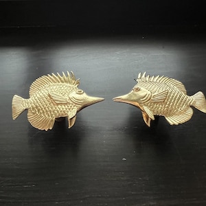 May include: Two gold-colored fish-shaped door knobs. The fish are facing each other with their mouths open.