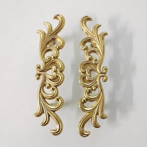 May include: Two ornate gold-toned metal drawer pulls with a decorative scroll design.