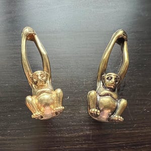 May include: Two gold monkey figurines with their arms raised above their heads. The monkeys are sitting on a dark brown surface.
