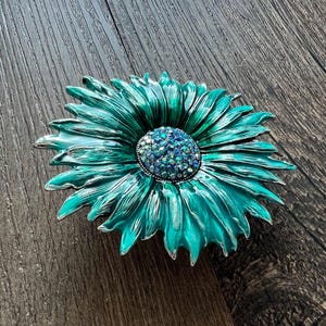 May include: A teal and silver flower-shaped brooch. The brooch has a central cluster of blue rhinestones surrounded by teal petals with silver edges. The brooch is a decorative accessory.