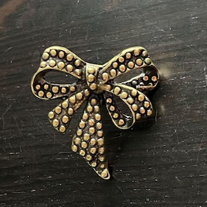 May include: A gold-toned bow-shaped brooch with a textured, dotted surface. The brooch has a classic bow design with two loops and two hanging tails. The metal has a slightly aged appearance, adding to its vintage charm. The brooch is a decorative accessory.