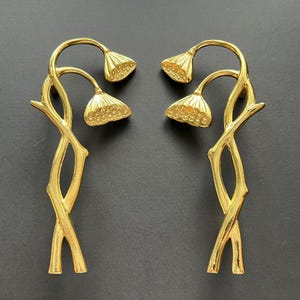 May include: Two gold-colored decorative floral sculptures. Each sculpture features a stem-like base with two curved branches, each holding a detailed, textured flower head. The pieces are symmetrical and set against a dark background.