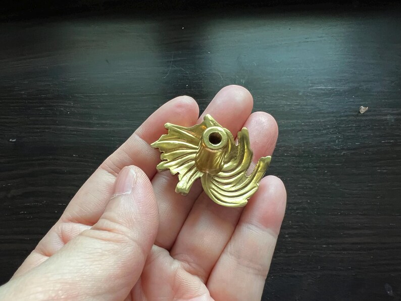 K&ouml;nnte beinhalten: Ein goldfarbener dekorativer Schrankknopf mit einem stilisierten, floralen Design. Der Knopf hat ein zentrales Loch und ein wirbelndes, blattartiges Muster. Die Hardware wird in einer Hand vor einem dunklen Hintergrund gehalten.