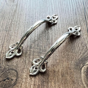 May include: Two silver cabinet handles with a decorative floral design. The handles are curved and have a polished finish, resting on a wooden surface. Suitable for furniture restoration or new builds.