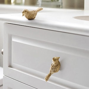 Puede incluir: Dos tiradores de caj&oacute;n en forma de p&aacute;jaro dorados en un armario blanco. El armario tiene un caj&oacute;n y un estante inferior.