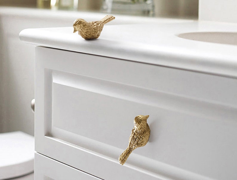 Puede incluir: Dos tiradores de caj&oacute;n en forma de p&aacute;jaro dorados en un armario blanco. El armario tiene un caj&oacute;n y un estante inferior.
