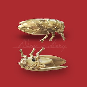 Peut inclure: Deux sculptures de cigales de couleur dorée. L'une est en position debout, les ailes déployées, l'autre est couchée sur le dos, les pattes en l'air.