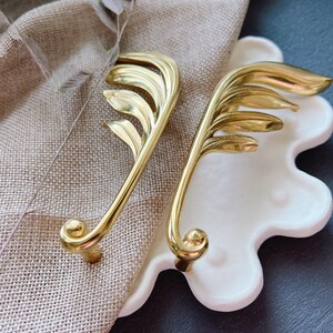 May include: Two gold leaf-shaped cabinet pulls with a decorative design. The pulls are on a white background.