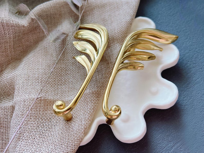 May include: Two gold leaf-shaped cabinet pulls with a decorative design. The pulls are on a white background.
