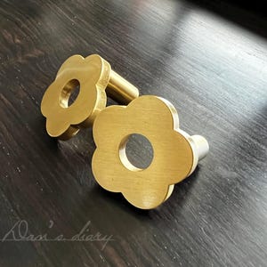 Pode incluir: Dois botões de metal de cor dourada em forma de flor com um furo no centro. Os botões estão sobre uma superfície de madeira escura.