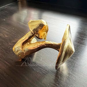 May include: A gold-colored metal sculpture of a person holding a large, flat, round object. The sculpture is abstract and stylized.