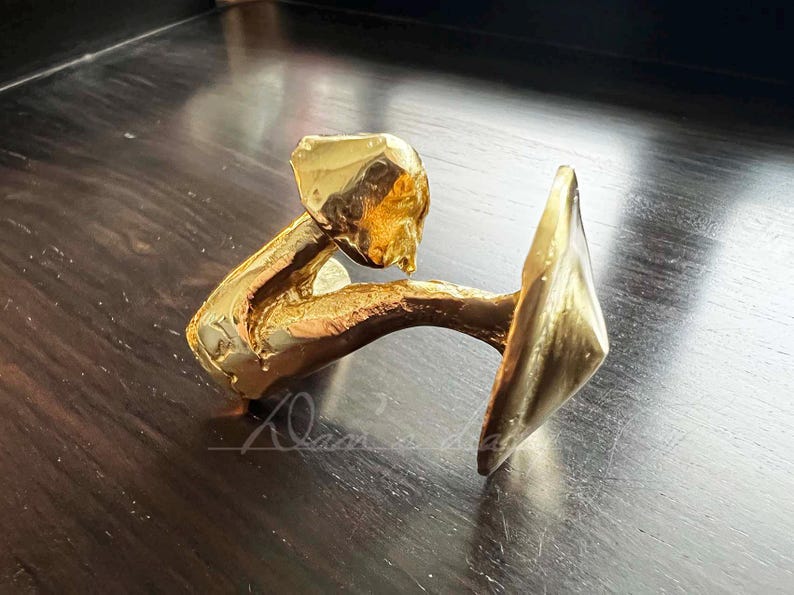 May include: A gold-colored metal sculpture of a person holding a large, flat, round object. The sculpture is abstract and stylized.