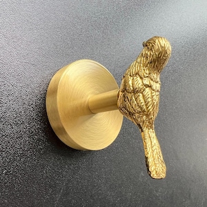 Op de afbeelding: Een goudkleurige vogelvormige wandhaak. De haak heeft een cirkelvormige basis en een staaf die zich uitstrekt naar de vogel. De vogel zit met gedetailleerde veren. De haak is gemonteerd op een zwarte, getextureerde ondergrond.