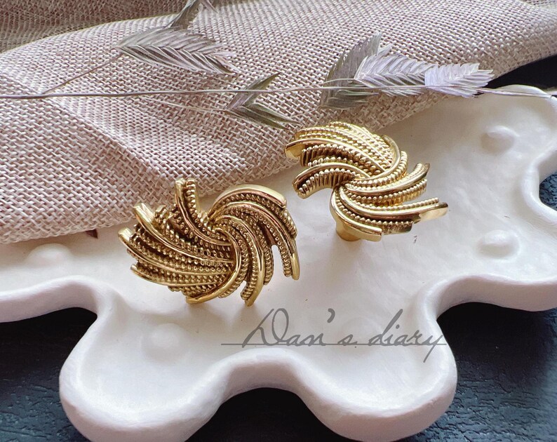 K&ouml;nnte beinhalten: Ein Paar goldfarbene, spiralf&ouml;rmige Ohrringe. Die Ohrringe haben ein strukturiertes, geriffeltes Design und werden auf einem wei&szlig;en, dekorativen Tablett pr&auml;sentiert. Das Tablett hat eine einzigartige, organische Form. Der Hintergrund umfasst einen beigen Stoff und dekorative Zweige.