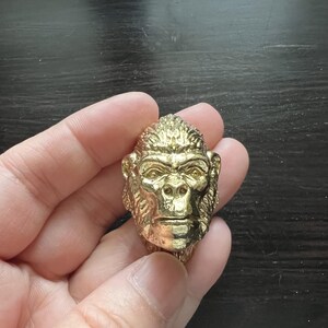May include: A gold-colored metal sculpture of a gorilla head. The sculpture is detailed and realistic, with a textured surface.
