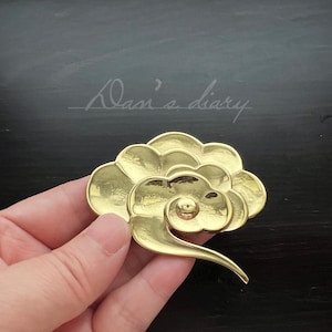 May include: A gold-colored metal brooch shaped like a stylized cloud with a swirl design. The brooch is a decorative accessory.