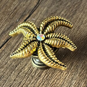 Op de afbeelding: Een goudkleurige kastknop in de vorm van een zeester met een centrale iriserende edelsteen. De knop heeft gedetailleerde ribbels en een bronskleurige basis, perfect voor een maritieme touch.