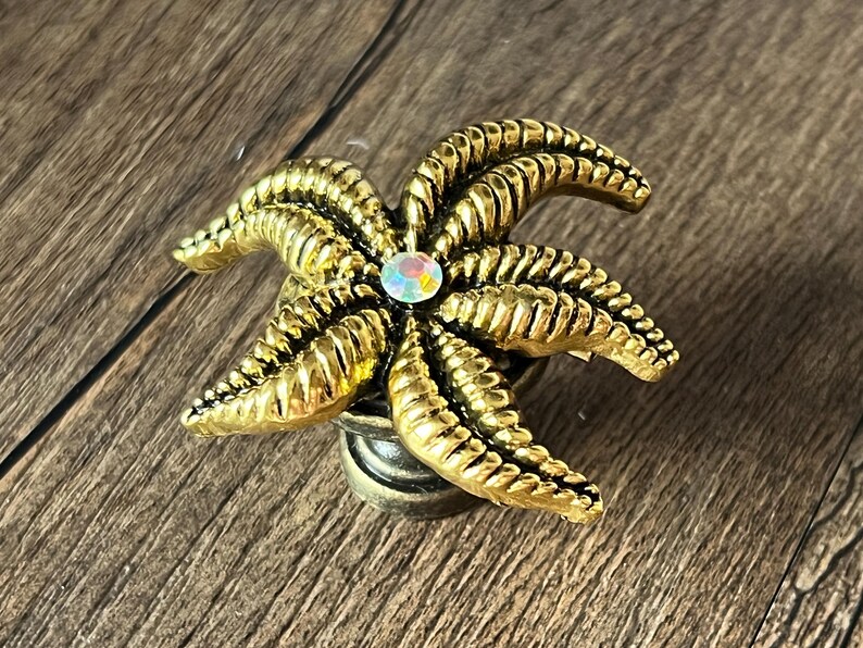 Op de afbeelding: Een goudkleurige kastknop in de vorm van een zeester met een centrale iriserende edelsteen. De knop heeft gedetailleerde ribbels en een bronskleurige basis, perfect voor een maritieme touch.