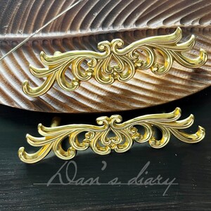 May include: Two ornate gold-toned metal drawer pulls with a decorative scroll design. The pulls are on a wooden surface.