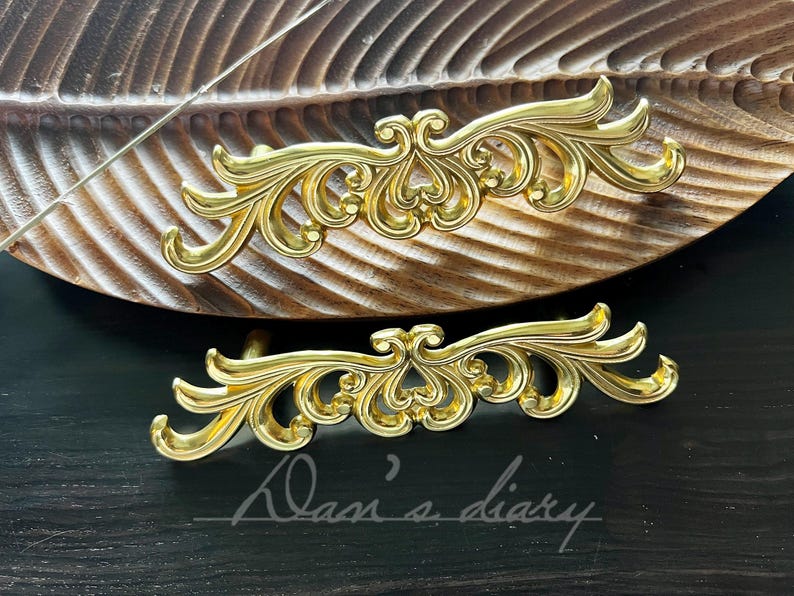 May include: Two ornate gold-toned metal drawer pulls with a decorative scroll design. The pulls are on a wooden surface.