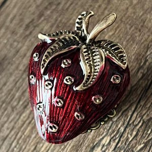 Op de afbeelding: Een gedetailleerde, rode aardbeivormige broche met goudkleurige accenten. De broche heeft een getextureerd oppervlak en kleine, goudkleurige stippen. De bladeren en de steel zijn ook goudkleurig, wat bijdraagt aan de charme van dit sieraad.
