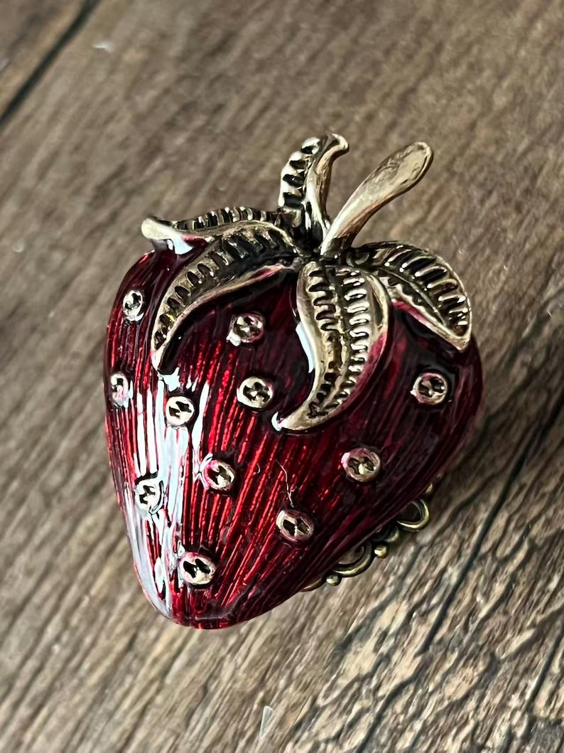 Op de afbeelding: Een gedetailleerde, rode aardbeivormige broche met goudkleurige accenten. De broche heeft een getextureerd oppervlak en kleine, goudkleurige stippen. De bladeren en de steel zijn ook goudkleurig, wat bijdraagt aan de charme van dit sieraad.