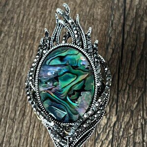 May include: An oval brooch featuring an abalone shell centrepiece, set within a silver-tone metal frame. The frame is adorned with small, dark grey rhinestones and a flame-like design. The abalone shell exhibits iridescent green and blue colours.