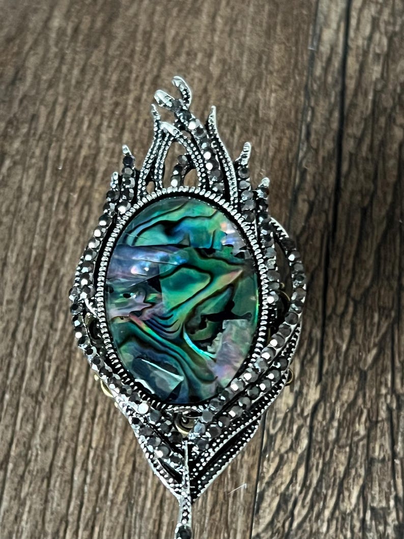 May include: An oval brooch featuring an abalone shell centrepiece, set within a silver-tone metal frame. The frame is adorned with small, dark grey rhinestones and a flame-like design. The abalone shell exhibits iridescent green and blue colours.