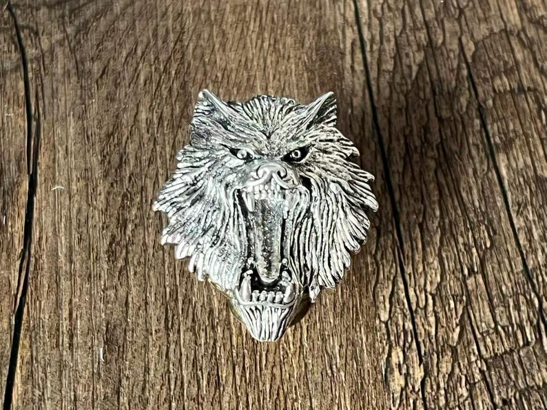 K&ouml;nnte beinhalten: Ein silberner Wolfskopf-Anh&auml;nger mit offenem Maul, der Z&auml;hne zeigt. Der Anh&auml;nger hat detailliertes Fell und ist vor einem Holzgrund platziert. Der Anh&auml;nger ist wahrscheinlich ein Schmuckst&uuml;ck.