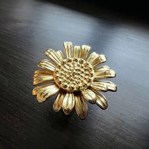 以下が含まれることがあります： ひまわり型の金色の金属製の引き出しの取っ手。ひまわりは、質感のある中心と花びらを持っています。