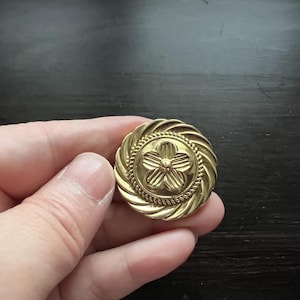May include: A gold-toned metal button with a raised flower design. The button has a rope-like border.