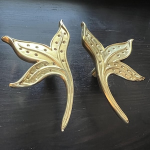 May include: Two brass-colored, leaf-shaped cabinet pulls. Each pull has a stylized floral design with a curved stem and a series of small, evenly spaced holes. The pulls are displayed against a dark background.