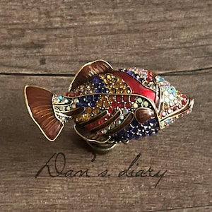 May include: A decorative fish figurine with a brown tail and fins, and a red stripe down the back. The fish is covered in colourful rhinestones in shades of blue, yellow, red, and green. The figurine is on a wooden surface.