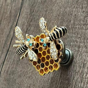 May include: A gold-toned bee-themed brooch featuring two bees with black and white striped bodies and crystal-like wings. The bees are positioned on a honeycomb with blue accents.