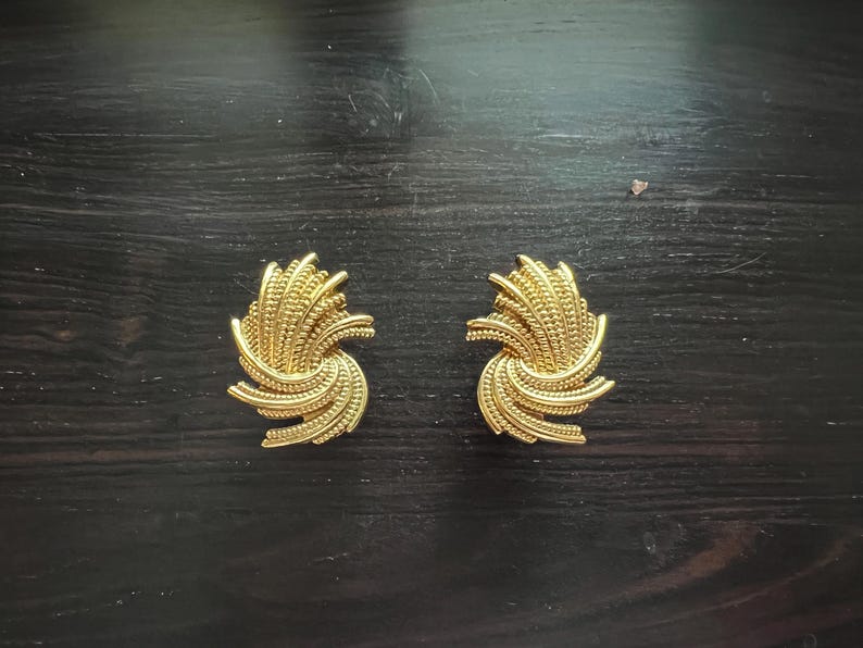 K&ouml;nnte beinhalten: Ein Paar goldfarbene Clip-Ohrringe. Jeder Ohrring hat ein wirbelndes, strukturiertes Design mit einem zentralen Cluster aus kleinen, perlenbesetzten Details. Die Ohrringe liegen auf einer dunklen Holzoberfl&auml;che.