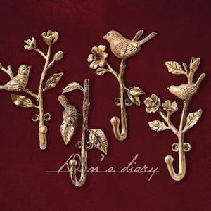 May include: Four decorative wall hooks in a gold finish, each featuring a bird perched on a branch with leaves and flowers. The hooks are mounted on a deep red background. The text "Aria's diary" is written in cursive.