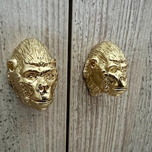 以下が含まれることがあります： 金色のゴリラの頭の形をしたキャビネットのドアノブが2つ。ゴリラの頭は前を向いていて、口は閉じている。