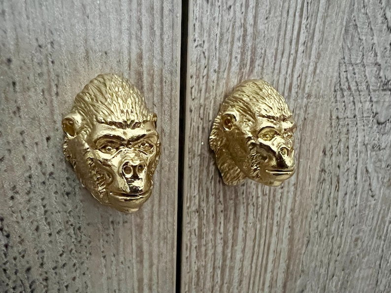 May include: Two gold-colored gorilla head cabinet door pulls. The gorilla heads are facing forward with their mouths closed.