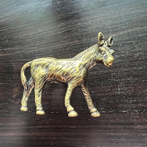 May include: A small, detailed brass donkey figurine. The donkey is standing with its legs spread, and its ears are perked up. The figurine has a textured surface and is set against a dark wooden background. A decorative item.